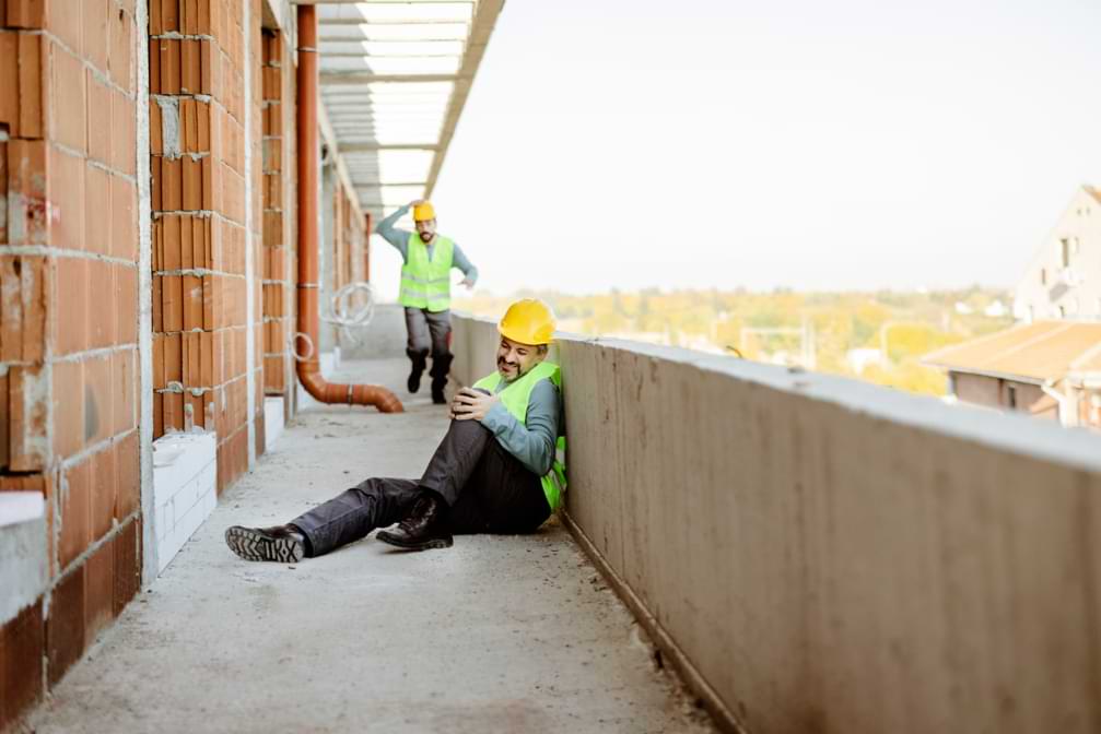 Injured construction worker siting on ground after Construction Sit accident with coworker running to help in background